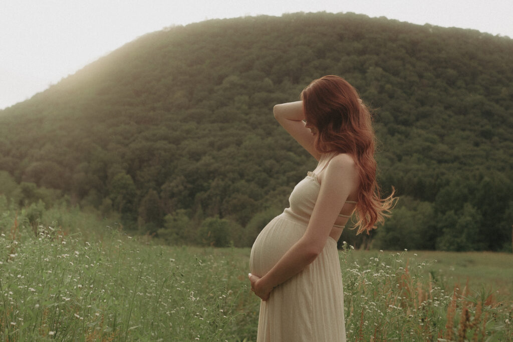 a woman stands in a green field with a hill in the distance holding her belly for maternity photos in a wildflower field