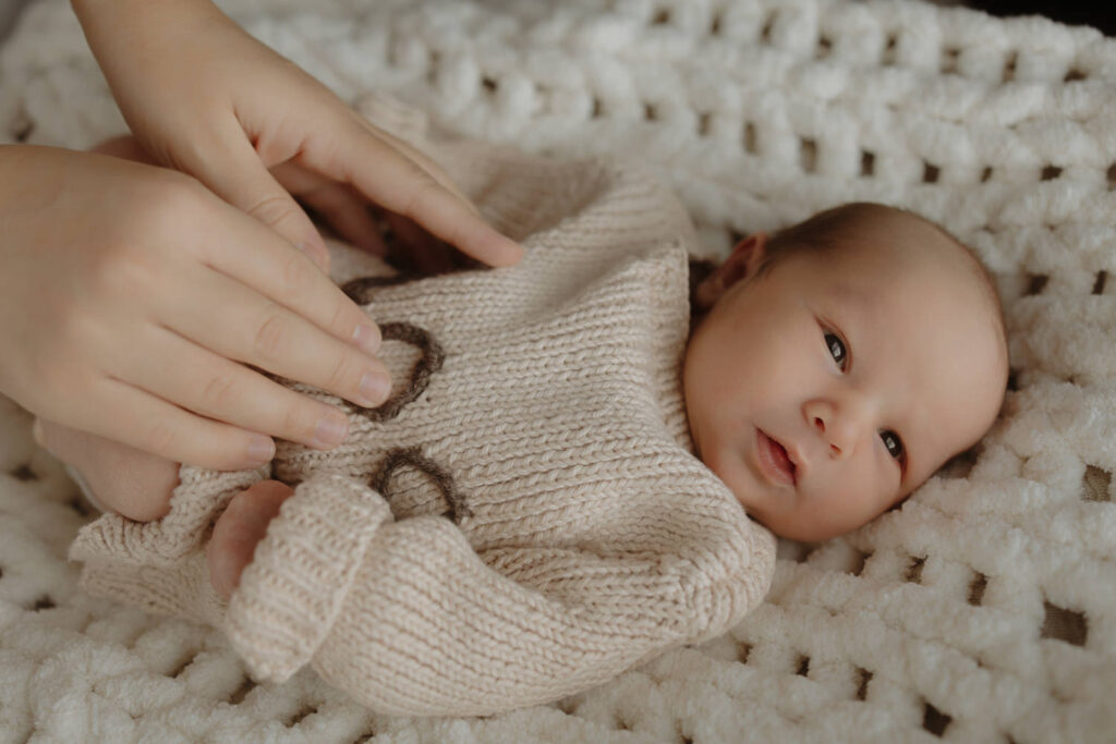 a newborn wearing a beige sweater and his mother's hands in a photo taken by a living room window at home